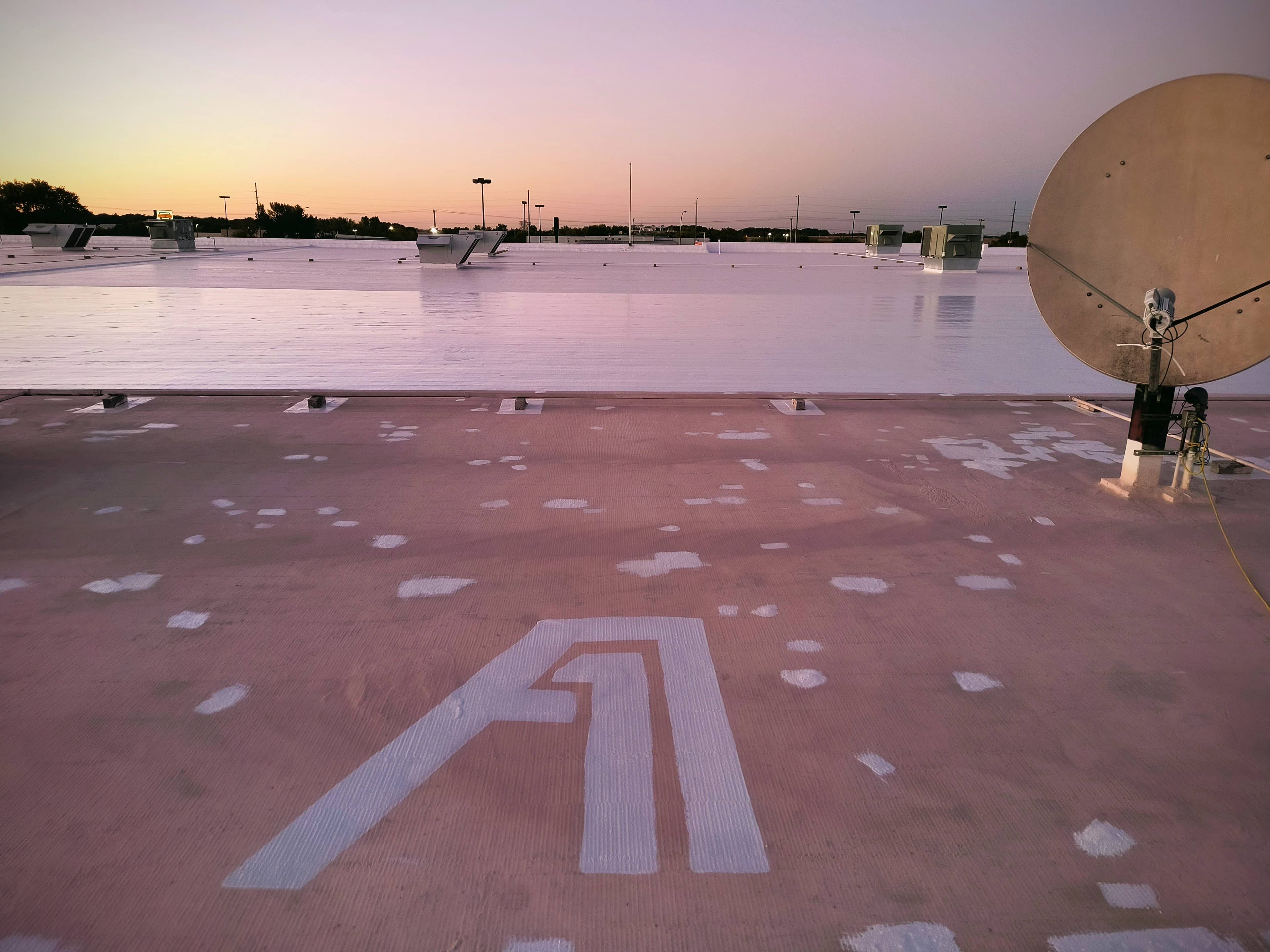 A commercial roof coating in progress at dusk: the finished reflective white topcoat across the far field, with the primed, patch-repaired substrate and A-1 Roofing's stencilled logo in the foreground